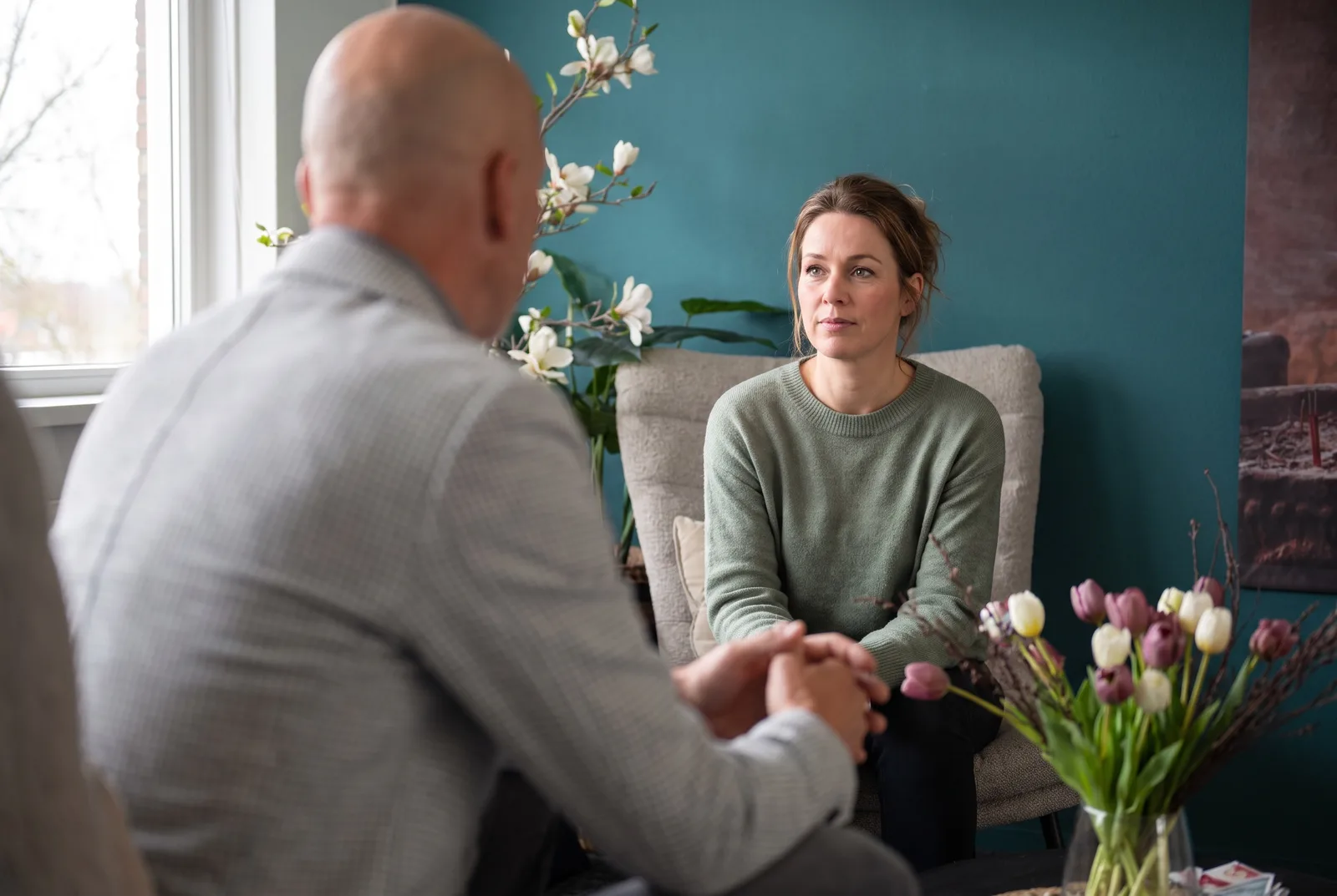 Coachgesprek onder vier ogen: over de schouder van de coach kijkt de coachee aandachtig en rustig terug, in de coachruimte met teal wand en magnolia
